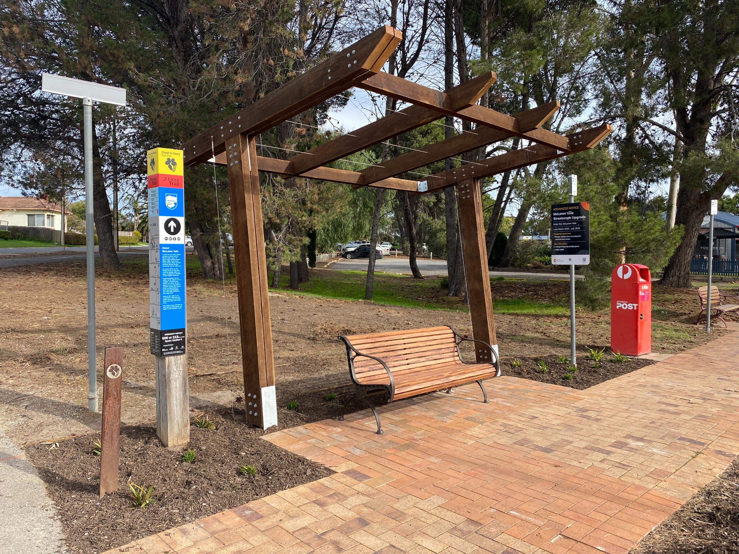 Timber bench under a wooden pergola near trail signage, a red Australia Post box, and a paved footpath in a tree-lined area.