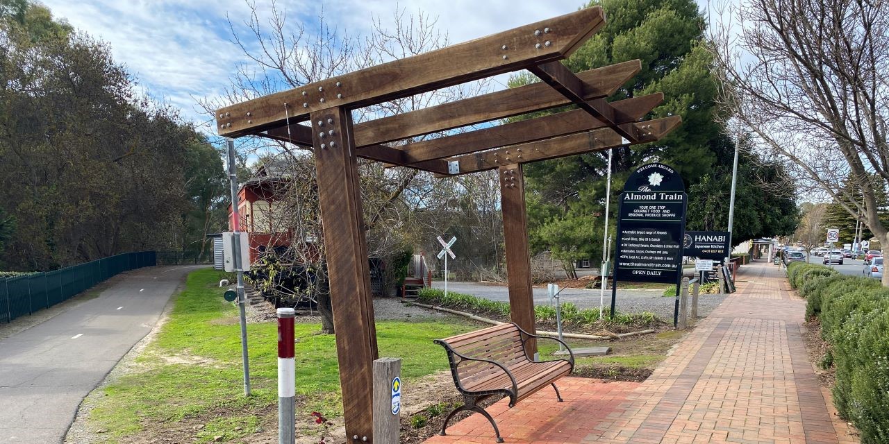 Wooden bench under a timber pergola structure next to a brick footpath near the Almond Train and bike trail entrance.