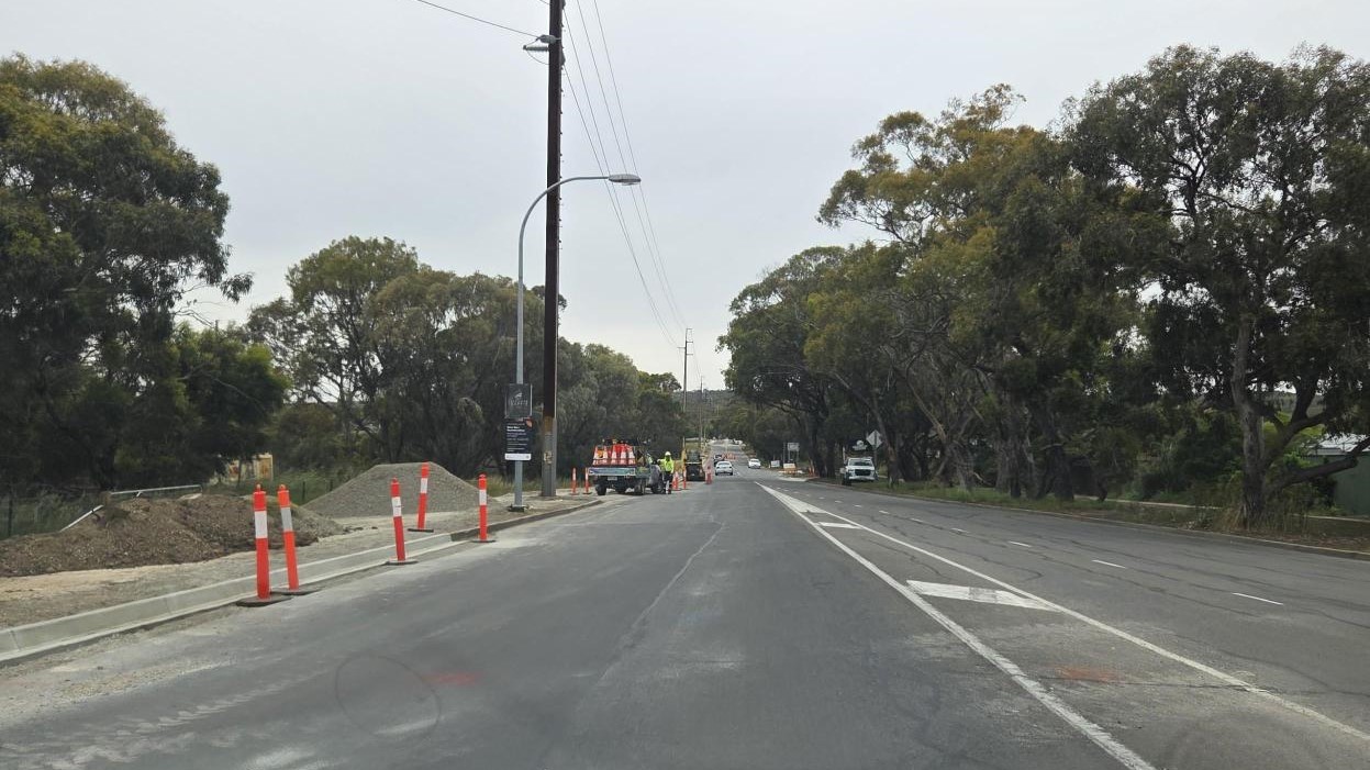 Roadworks in progress along Bains Road, with construction workers visible ahead and temporary orange bollards placed along the left side of the road. New kerb and gutter sections are partially completed, and trees line both sides of the street under a clo