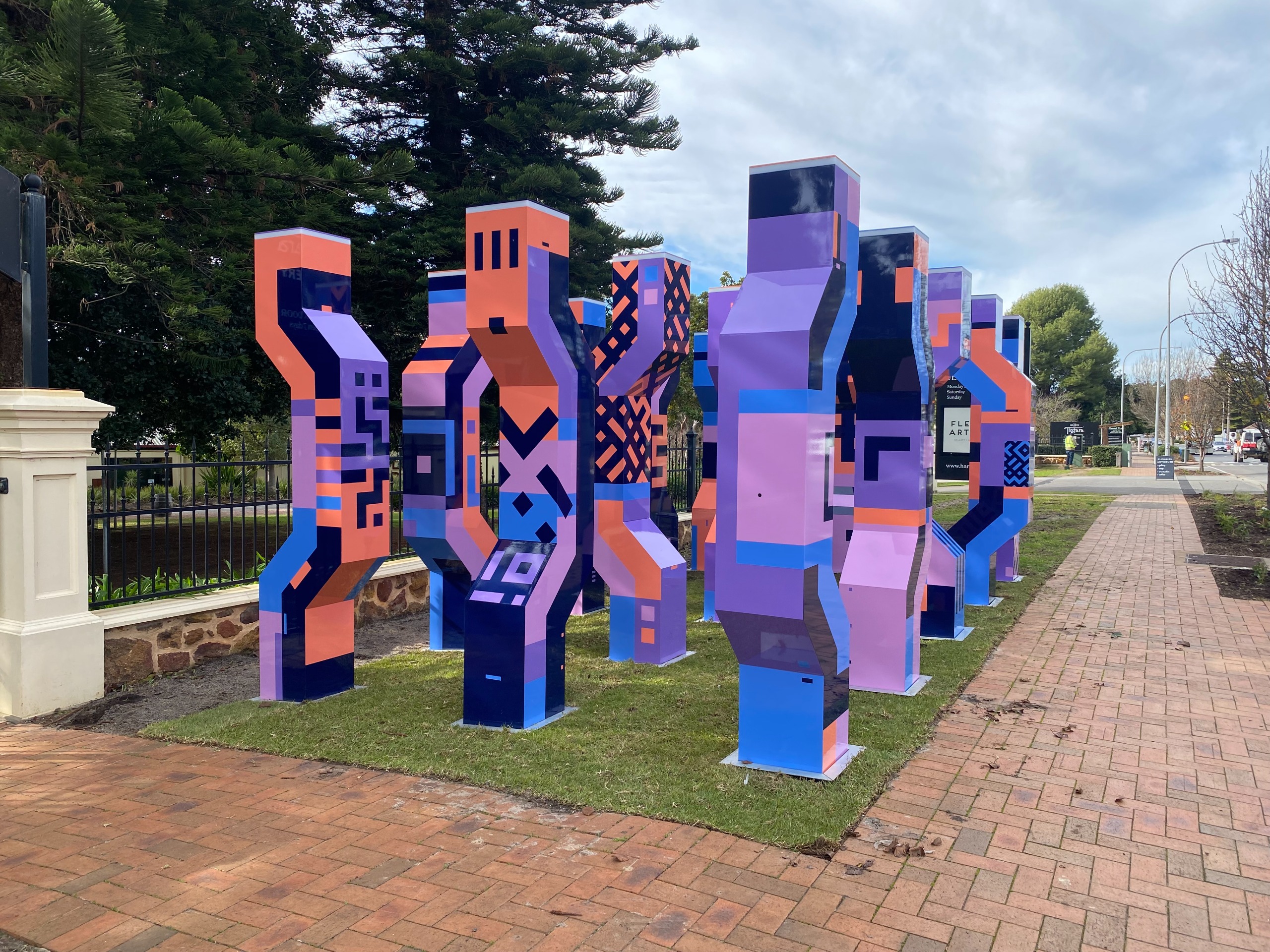 Brightly coloured geometric pillars arranged on grass near a paved footpath and fence, with trees and street visible in the background.